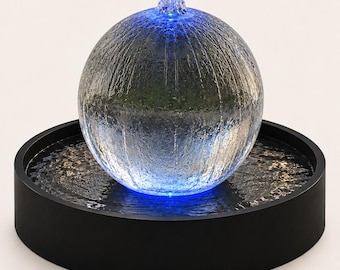 Könnte beinhalten: Ein kugelförmiger Zimmerbrunnen mit einer klaren, texturierten Glaskugel, die in einem schwarzen, kreisförmigen Becken ruht. Der Boden der Glaskugel ist mit einem hellen blauen Licht beleuchtet, das einen leuchtenden Effekt auf der Wasseroberfläche erzeugt.
