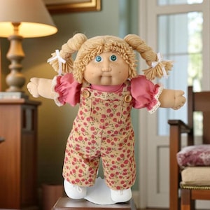 May include: A vintage doll with blonde pigtails tied with white ribbons. The doll has blue eyes, freckles, and is wearing a pink shirt with ruffled sleeves and a floral print overall. The doll is standing on a white stand.