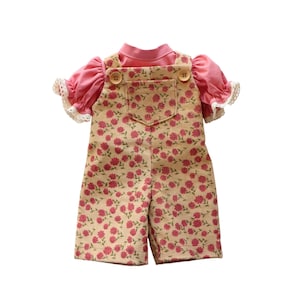 May include: A doll's outfit comprising a pink top with frilled sleeves and a beige floral-patterned overall. The overall has wooden buttons and a front pocket. The outfit is designed for a doll.