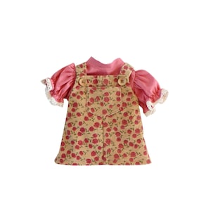 May include: A doll's outfit comprising a pink top with ruffled sleeves and a beige corduroy pinafore dress. The dress is patterned with red flowers and has wooden button fastenings. The sleeves have white lace trim.