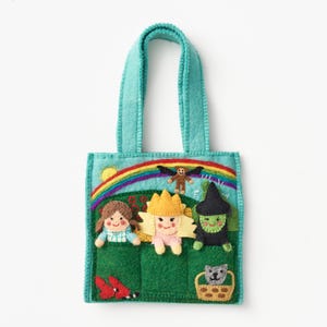 May include: A teal felt tote bag featuring an embroidered scene of a rainbow, a yellow sun, and a flying monkey. The bag displays three felt figures: a girl in a blue dress, a blonde character in a crown, and a green witch, with a cat in a basket.