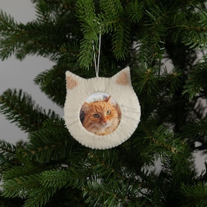 May include: A cream-colored cat-shaped Christmas ornament with a photo of an orange tabby cat in the center. The ornament has tan inner ears and white whiskers. It hangs from a white string against a green Christmas tree.