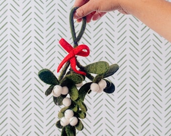 May include: A felted mistletoe ornament featuring dark green leaves and clusters of small white berries. The ornament is tied with a bright red felt bow and includes a green felt loop at the top for hanging, crafted from soft textile material.