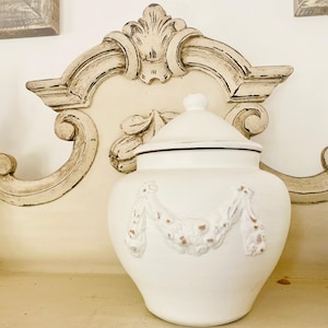 Peut inclure: Pot en céramique blanc avec couvercle, orné d'un motif en relief. Le pot a une forme arrondie et est posé sur une surface claire. L'arrière-plan comprend une tête de lit décorative en bois.