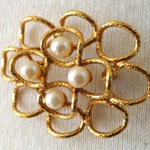 May include: A gold-toned brooch featuring an openwork, textured metal frame composed of interconnected, rounded loops. Four lustrous, cream-colored spherical pearls are set within the center of the metal structure, creating a floral-like arrangement.