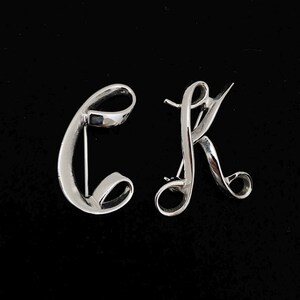 May include: Two silver-toned brooches in the shape of the letters "C" and "K" in a cursive script. The brooches have a polished finish and are set against a black background. The "C" brooch is on the left, and the "K" is on the right.