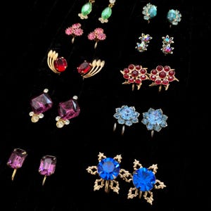 May include: A collection of ten pairs of vintage clip-on earrings featuring various colorful rhinestones, including red, blue, purple, and green stones set in gold and silver-toned metal frames, arranged in rows on a black velvet display surface.