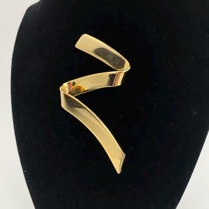 May include: A gold-colored metal ribbon brooch with a wavy design.