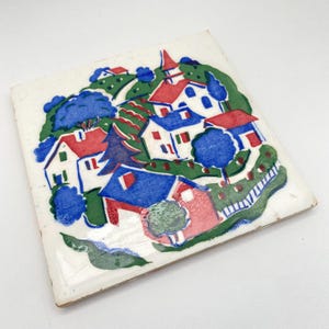 May include: A white ceramic tile with a hand-painted design of a small village with red, blue, and green houses, trees, and a winding road.