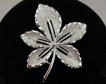 May include: A silver-toned brooch in the shape of a leaf. The leaf has a textured surface and is detailed with intricate cutouts.