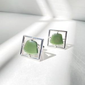 May include: A pair of square silver cufflinks, each featuring a light green, rounded rectangular stone set within a square frame. The cufflinks are displayed on a white surface, with soft shadows.