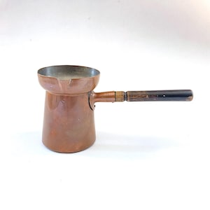 May include: A copper coffee pot with a wooden handle. The pot has a rounded base and a flared top. The handle is dark brown and black. The pot is likely used for making Turkish or Greek coffee.