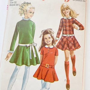 May include: A sewing pattern envelope featuring illustrations of three dresses for a size 10 girl with a 28.5 inch breast measurement. The designs include a long-sleeved green dress with a white sash, a short-sleeved orange dress with a belt, and a plaid dress.