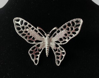 May include: A silver butterfly brooch with a delicate, intricate design. The wings are open and the body is detailed with a textured finish.