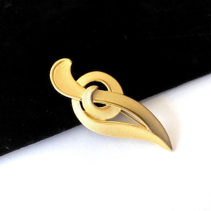 May include: A gold-toned brooch with an abstract design. The brooch features a curved, leaf-like shape connected to a circular element. The metal has a matte finish, and the brooch is set against a black background.