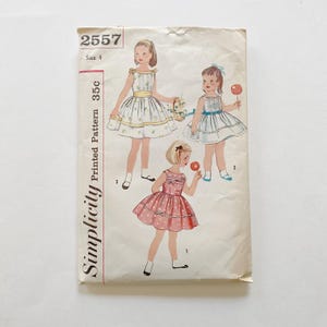 May include: Vintage Simplicity sewing pattern 2557, size 4. The cover features illustrations of three young girls in dresses. The dresses are in white, pink, and blue, with floral and polka dot patterns. The pattern is printed on aged paper.