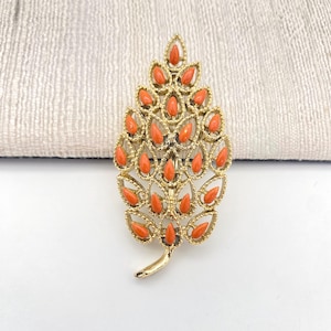 May include: A gold-toned leaf-shaped brooch with orange-red accents. The brooch features a detailed design with small, teardrop-shaped orange stones set within a gold filigree frame. The brooch has a curved stem at the base.