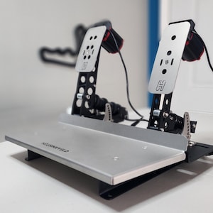 May include: Silver and black racing simulator pedal set. The set includes two pedals with a silver base and black accents. The pedals are connected by black wires. The base has the text "HEUSINKVELD" printed on it.
