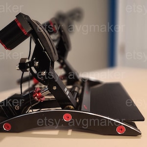 Peut inclure: Un ensemble de pédales de simulateur de course noir et rouge. L'ensemble comprend une base noire avec des accents et du matériel rouges. Les pédales sont inclinées et semblent réglables. L'image est prise d'en bas.