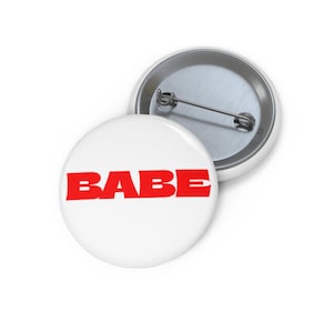 Może przedstawiać: Biała przypinka z napisem "BABE" pogrubioną czerwoną czcionką. Przypinka jest okrągła i ma metalowy tył z zapięciem na agrafkę. Prosty, ale przyciągający wzrok dodatek.