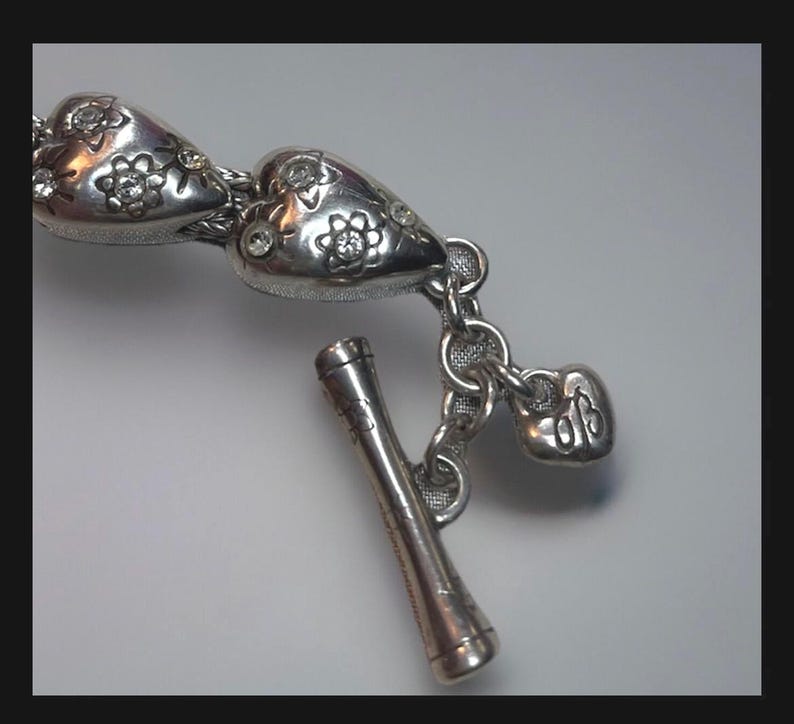 Peut inclure: Bracelet en argent avec deux breloques en forme de c&oelig;ur, chacune orn&eacute;e d'un motif floral et de petits strass. Le bracelet est dot&eacute; d'un fermoir &agrave; glissi&egrave;re avec un petit pendentif en forme de c&oelig;ur sur lequel est inscrit "925".