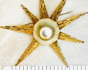 May include: A gold-toned starburst brooch featuring a central round cream-colored faux pearl. The metal rays have a textured, rough finish. A ruler placed at the bottom indicates the brooch measures approximately 2.5 inches in total width.