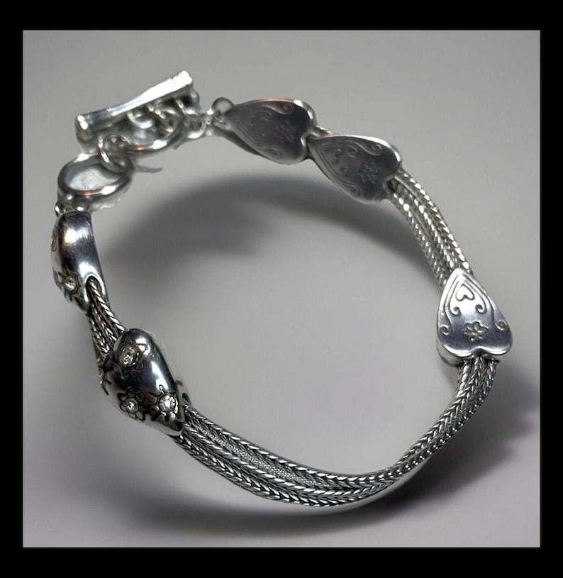 Peut inclure: Bracelet en argent avec un design de cha&icirc;ne et des breloques en forme de c&oelig;ur. Les breloques ont des d&eacute;tails complexes et certaines ont de petites pierres claires.