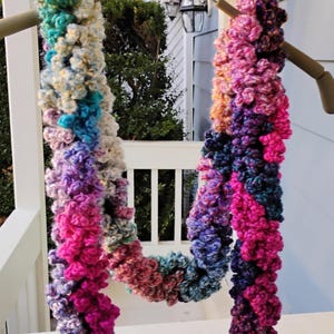 May include: A handmade, colorful scarf with a textured, looped design. The scarf features a gradient of colors, including pink, purple, blue, and green, and is displayed on a wooden hanger. The scarf is made of a soft, fluffy yarn.