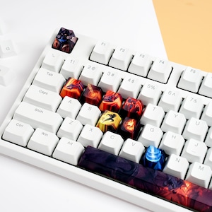 Może przedstawiać: Biała klawiatura mechaniczna z niestandardowymi nakładkami na klawisze. Nakładki na klawisze mają kolorowe wzory z odcieniami czerwieni, pomarańczy i niebieskiego. Niektóre nakładki są rozrzucone wokół klawiatury.