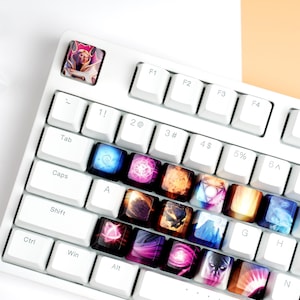 Puede incluir: Teclado mecánico blanco con teclas personalizadas con arte de fantasía colorido. Las teclas muestran varios diseños, incluyendo orbes brillantes y retratos de personajes. Teclas blancas adicionales están dispersas.