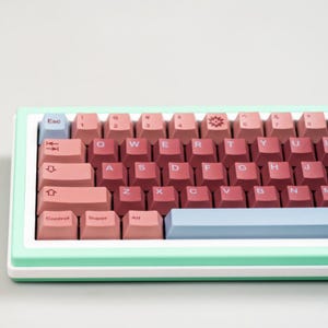 May include: A pastel-colored mechanical keyboard with a mint green frame and white base. The keycaps are a gradient of pink and red, with a light blue escape key and space bar. The keyboard has a standard QWERTY layout.