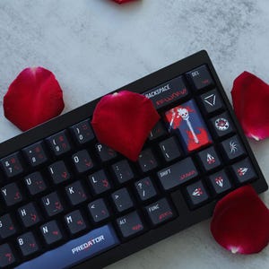 May include: Black mechanical keyboard with custom keycaps, including a Predator key and a keycap with a skull and sword design. Red rose petals are scattered around the keyboard.