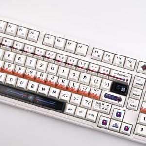 May include: White mechanical keyboard with custom keycaps. The keycaps feature a mix of white, black, and red designs, including pixelated graphics and text. The keyboard has a standard layout with function keys and a number pad.