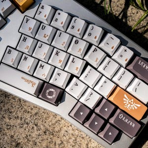 Puede incluir: Un teclado mecánico personalizado con teclas blancas y grises. Las teclas tienen letras y símbolos dorados y marrones. Una tecla es naranja con un emblema blanco. El teclado está sobre una base gris.