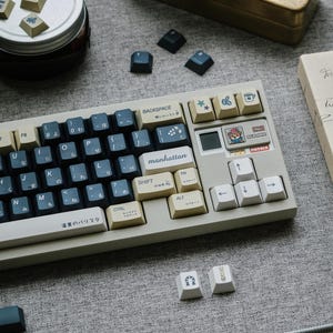 Op de afbeelding: Een aangepast mechanisch toetsenbord met een lichtgrijze basis, zwarte en crèmekleurige toetsen en decoratieve toetsen. Het toetsenbord heeft een compacte indeling en heeft een "manhattan"-toets. Extra toetsen en een notitieboek zijn ook zichtbaar.