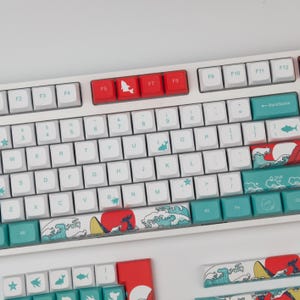 May include: White mechanical keyboard with custom keycaps. The keycaps feature a Japanese wave design with red accents and fish icons. The function keys are red. The keyboard has a standard QWERTY layout.
