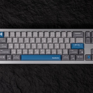 May include: A gray and blue mechanical keyboard with white keycaps. The keyboard has a blue space bar with the text "NoodlesSort". The keyboard also has a blue "ON CALL" key and a "404" key.
