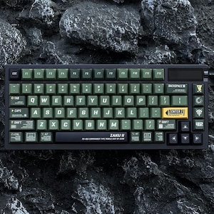 Puede incluir: Un teclado mecánico negro con teclas verde oliva y letras blancas. El teclado tiene forma rectangular con un marco negro y una tecla "ENTER" amarilla. El texto "ZAKU II" está impreso en la parte inferior.