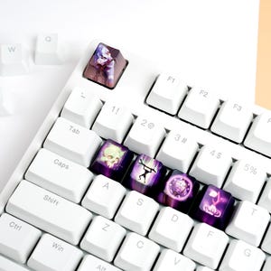 Op de afbeelding: Wit mechanisch toetsenbord met aangepaste keycaps. De keycaps tonen verschillende ontwerpen in paars, roze en groen. Sommige keycaps zijn verwijderd en verspreid over het witte oppervlak.