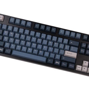 May include: A black mechanical keyboard with blue and gray keycaps. The keyboard has a standard layout with function keys, arrow keys, and a number pad. The keycaps are a mix of blue and gray, with white lettering. The keyboard is angled towards the user.