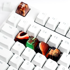 Puede incluir: Un teclado mecánico blanco con teclas personalizadas. Las teclas son coloridas, con diseños en tonos marrones, rojos, verdes y naranjas. Las teclas del teclado están dispuestas en una distribución QWERTY estándar.