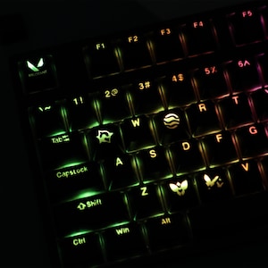 May include: A black mechanical keyboard with customizable backlit keys featuring a gradient of green, yellow, and red light. Several keys display unique white icons, including the Valorant logo, a wave, and butterfly shapes, replacing standard letters.