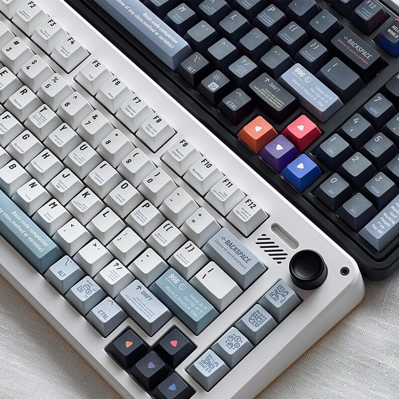 May include: Two mechanical keyboards are shown, one white and one black. The white keyboard has white and grey keycaps, while the black keyboard has black and grey keycaps. Both keyboards feature function keys and a volume control.