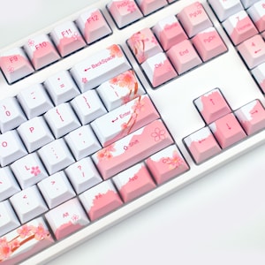 Puede incluir: Un teclado mecánico blanco con teclas rosas. Las teclas presentan diseños y acentos de flores de cerezo. El teclado tiene teclas estándar, incluidas teclas de función, teclas de flecha y un teclado numérico. Las teclas son una mezcla de blanco y rosa.
