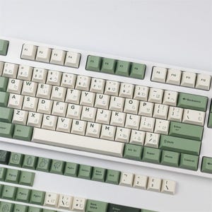 Könnte beinhalten: Eine weiße mechanische Tastatur mit grünen und cremefarbenen Tastenkappen. Die Tastatur hat japanische Zeichen und englische Buchstaben. Zusätzliche Tastenkappen sind enthalten, mit Kaktus- und Blattmotiven.