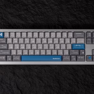 May include: A mechanical keyboard with gray keycaps and a silver frame. The keyboard has blue accent keys, including the space bar with "NoodleSort" printed on it. The keyboard also has the text "ON CALL" and "404" on some keys.