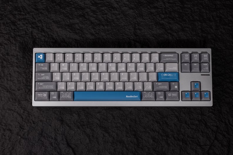 May include: A mechanical keyboard with gray keycaps and a silver frame. The keyboard has blue accent keys, including the space bar with "NoodleSort" printed on it. The keyboard also has the text "ON CALL" and "404" on some keys.