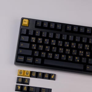 May include: A black mechanical keyboard with gold lettering and a gold "Esc" key. Additional keycaps with hieroglyphic designs are displayed below the keyboard. The keyboard has a standard QWERTY layout.