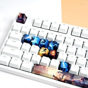 Könnte beinhalten: Eine weiße mechanische Tastatur mit individuellen Tastenkappen. Die Tastenkappen weisen verschiedene Designs mit blauen, goldenen und roten Akzenten auf. Einige Tastenkappen sind um die Tastatur verstreut.