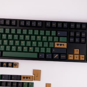 May include: A black mechanical keyboard with green and gold keycaps. The keys are arranged in a standard QWERTY layout. Some keycaps feature heart symbols and a sword icon. The keyboard is on a white surface.